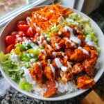 A colorful buffalo chicken bowl featuring orange buffalo chicken pieces over rice, topped with ranch dressing, shredded carrots, lettuce, and cherry tomatoes.